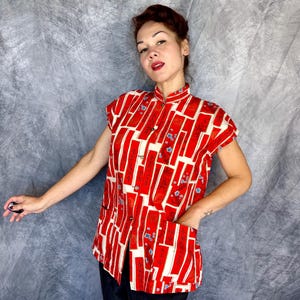 Pode incluir: Uma camisa de manga curta com bot&otilde;es, apresentando um padr&atilde;o geom&eacute;trico vibrante em vermelho e creme com blocos retangulares e pequenos detalhes florais azuis. A camisa tem gola mandarim, fecho frontal de bot&otilde;es e dois bolsos grandes aplicados.