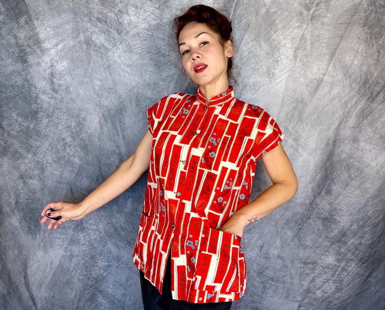 Pode incluir: Uma camisa de manga curta com bot&otilde;es, apresentando um padr&atilde;o geom&eacute;trico vibrante em vermelho e creme com blocos retangulares e pequenos detalhes florais azuis. A camisa tem gola mandarim, fecho frontal de bot&otilde;es e dois bolsos grandes aplicados.