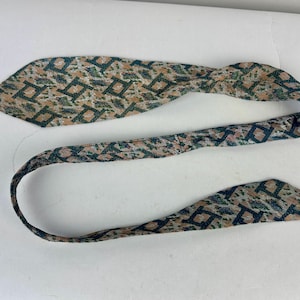 May include: A necktie featuring a repeating geometric pattern of dark blue squares on a textured background of tan, beige, and muted green. The fabric has a woven appearance with a pointed end and a long, narrow strap that curves across the frame.