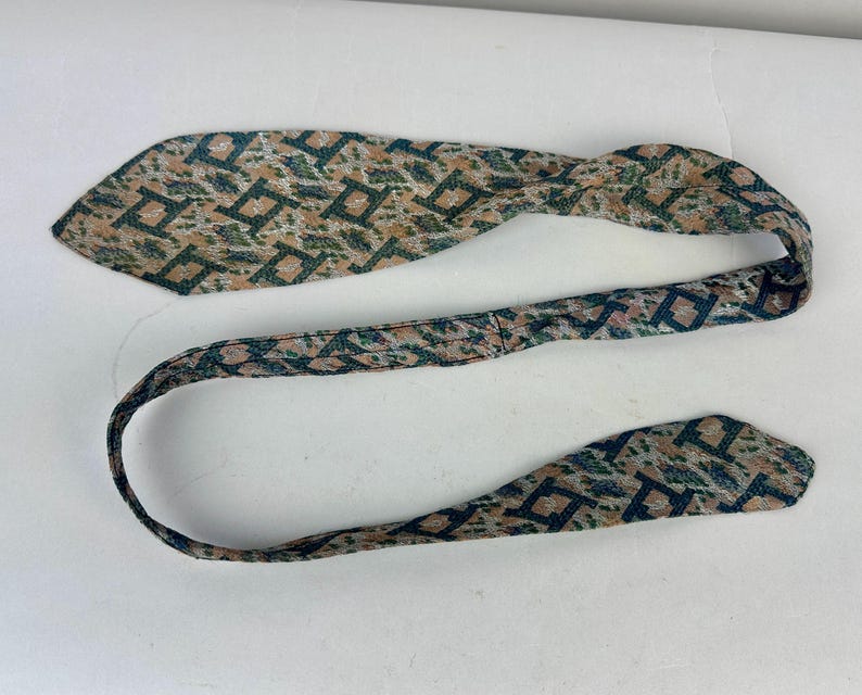 May include: A necktie featuring a repeating geometric pattern of dark blue squares on a textured background of tan, beige, and muted green. The fabric has a woven appearance with a pointed end and a long, narrow strap that curves across the frame.