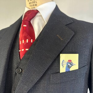 May include: A dark charcoal gray pinstriped suit jacket and vest worn over a crisp white collared shirt. A vibrant red necktie with a geometric white pattern is tied at the neck. A light yellow pocket square features a peacock wearing a red hat.