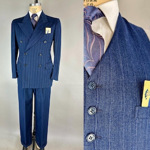 Puede incluir: Un traje azul marino de doble botonadura con sutiles rayas diplomáticas. El traje incluye una chaqueta, un pantalón y un chaleco. Una corbata estampada y un pañuelo de bolsillo añaden detalles. El traje se exhibe en un maniquí.
