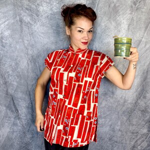 Pode incluir: Uma camisa de manga curta com gola mandarim, apresentando um padr&atilde;o geom&eacute;trico vibrante em vermelho e creme com pequenos detalhes florais azuis. A camisa tem um fecho frontal de bot&otilde;es e &eacute; acompanhada por uma caneca cil&iacute;ndrica de cer&acirc;mica verde-oliva.