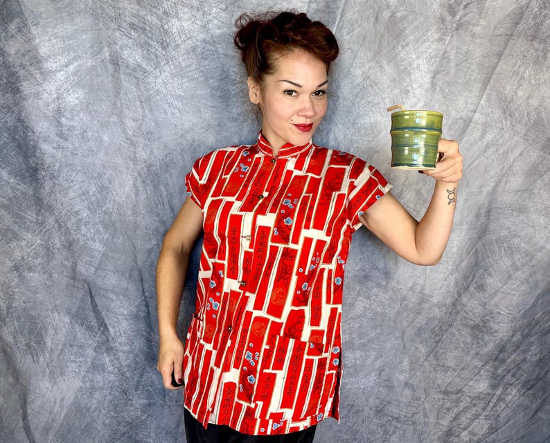 Pode incluir: Uma camisa de manga curta com gola mandarim, apresentando um padr&atilde;o geom&eacute;trico vibrante em vermelho e creme com pequenos detalhes florais azuis. A camisa tem um fecho frontal de bot&otilde;es e &eacute; acompanhada por uma caneca cil&iacute;ndrica de cer&acirc;mica verde-oliva.