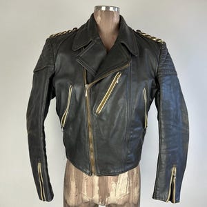 Puede incluir: Una chaqueta de moto de cuero negro con detalles de cremallera dorada y cuello con cremallera. La chaqueta tiene un diseño acolchado en los hombros y los codos.