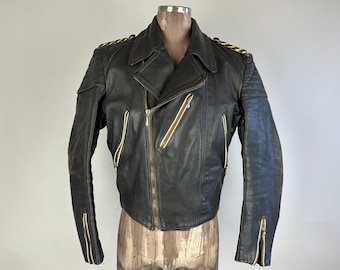 Può includere: Una giacca da motociclista in pelle nera con dettagli in zip dorati e un colletto con zip. La giacca ha un design trapuntato sulle spalle e sui gomiti.