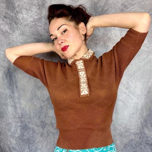 Pu&ograve; includere: Un maglione marrone a maniche corte con colletto decorativo. Il maglione ha una trama a coste e un motivo geometrico sul davanti. Il colletto presenta un design floreale color crema e beige.