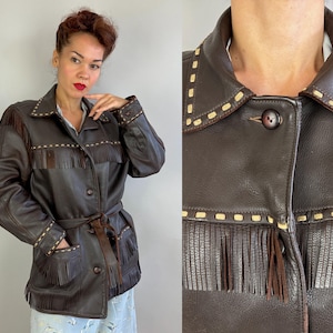 May include: A dark brown leather jacket with fringe detailing on the chest, pockets, and sleeves. The jacket has a button-up front, a collar, and a matching belt. The stitching is a lighter brown color.