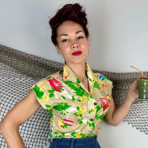 May include: Tan short-sleeved button-up shirt with a tropical print featuring red birds, cocktails, and flowers. The shirt has a classic collar and is paired with blue denim bottoms. A green ceramic cup with a straw is held.