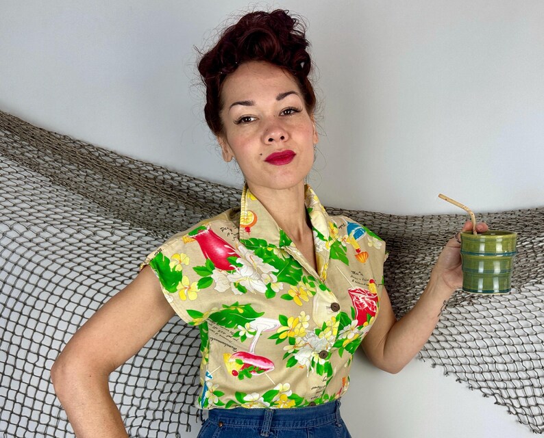 May include: Tan short-sleeved button-up shirt with a tropical print featuring red birds, cocktails, and flowers. The shirt has a classic collar and is paired with blue denim bottoms. A green ceramic cup with a straw is held.