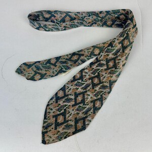 May include: A necktie featuring a woven geometric pattern in shades of dark green, tan, and muted blue. The fabric displays a repeating angular design across the entire surface, with a pointed end and a folded structure that shows the interior lining.