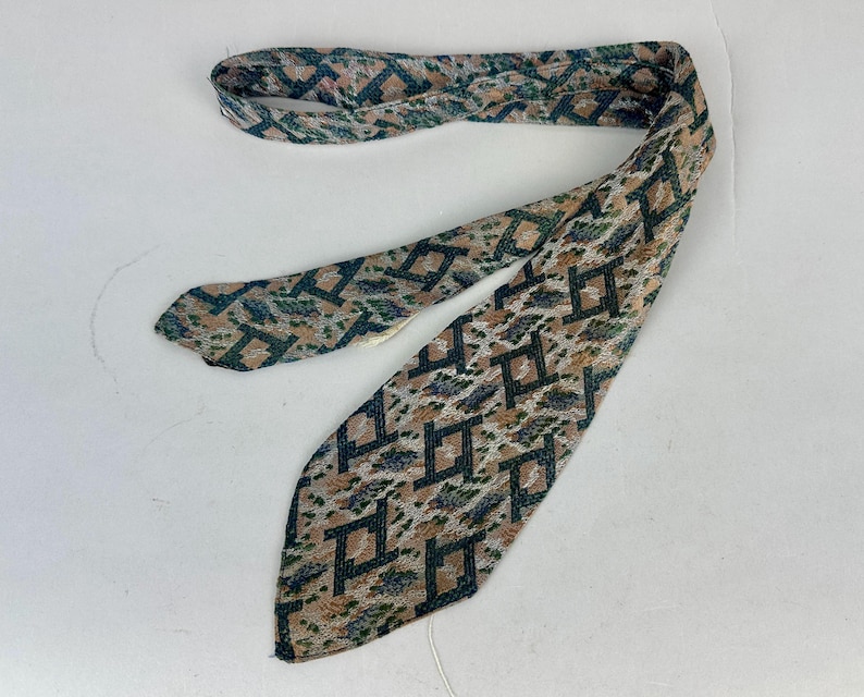 May include: A necktie featuring a woven geometric pattern in shades of dark green, tan, and muted blue. The fabric displays a repeating angular design across the entire surface, with a pointed end and a folded structure that shows the interior lining.