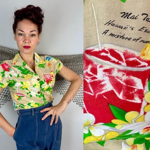 May include: Tan short-sleeved button-up shirt with a tropical print featuring a cocktail, flowers, and flamingos. The shirt has a collared neckline and wooden buttons. The text on the shirt reads "Mai Tai Hawaii's Exotic A mixture of rums."