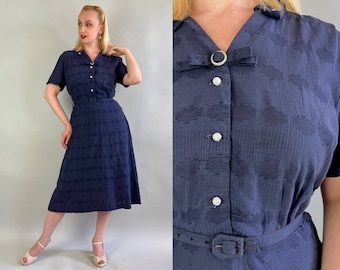 May include: A navy blue dress with a white button-up front and a white belt. The dress has a textured pattern and a short sleeve.