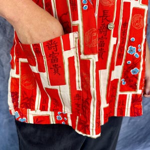 Pode incluir: Uma camisa vermelha de bot&otilde;es com um padr&atilde;o repetitivo de pain&eacute;is retangulares verticais com caracteres kanji pretos, motivos florais circulares e pequenas flores azuis. A camisa inclui um bolso frontal e bot&otilde;es dourados.