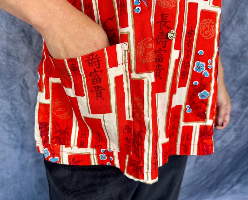 Pode incluir: Uma camisa vermelha de bot&otilde;es com um padr&atilde;o repetitivo de pain&eacute;is retangulares verticais com caracteres kanji pretos, motivos florais circulares e pequenas flores azuis. A camisa inclui um bolso frontal e bot&otilde;es dourados.