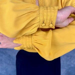 May include: Mustard yellow blouse with gathered sleeves and buttoned cuffs. The cuffs have a textured, grid-like pattern. The blouse is paired with a navy blue garment, creating a contrast in color.