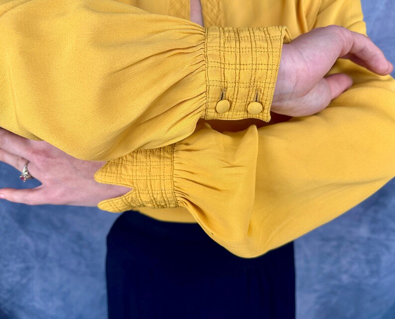 May include: Mustard yellow blouse with gathered sleeves and buttoned cuffs. The cuffs have a textured, grid-like pattern. The blouse is paired with a navy blue garment, creating a contrast in color.