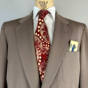 May include: A taupe suit jacket with a white dress shirt and a patterned tie. The tie features a red and cream design. A small card with a peacock illustration is in the breast pocket. The suit is displayed on a mannequin.