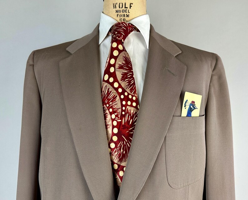 May include: A taupe suit jacket with a white dress shirt and a patterned tie. The tie features a red and cream design. A small card with a peacock illustration is in the breast pocket. The suit is displayed on a mannequin.