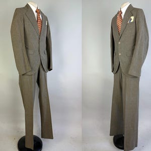 Op de afbeelding: Een tweedelig pak in een gedempte olijfbruine tint, bestaande uit een single-breasted colbert met twee knopen, inkepingen in de revers en klepzakken. Het pak bevat een bijpassende broek, een wit overhemd met kraag en een das met patroon.