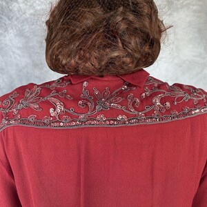 May include: A burgundy blouse featuring detailed sequin and bead embroidery across the shoulders. The embellishments create a floral design along the collar and shoulder. The blouse has a traditional collar and a smooth, flowing fabric.