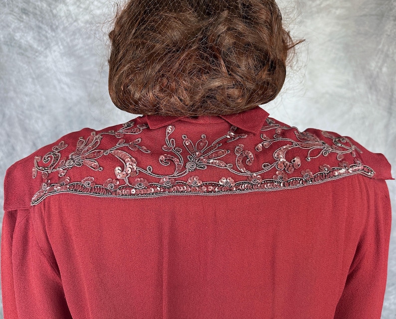 May include: A burgundy blouse featuring detailed sequin and bead embroidery across the shoulders. The embellishments create a floral design along the collar and shoulder. The blouse has a traditional collar and a smooth, flowing fabric.