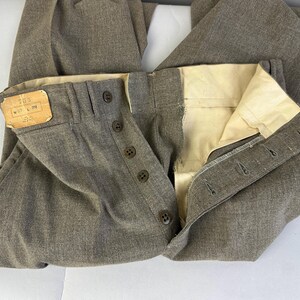 Op de afbeelding: Taupekleurige broek met een knoopsluiting met vijf donkerbruine knopen. De binnenkant van de tailleband is gevoerd met cr&egrave;mekleurige stof. Een rechthoekig beige label aan de linkerkant toont de tekst 755, W 37 L 32.