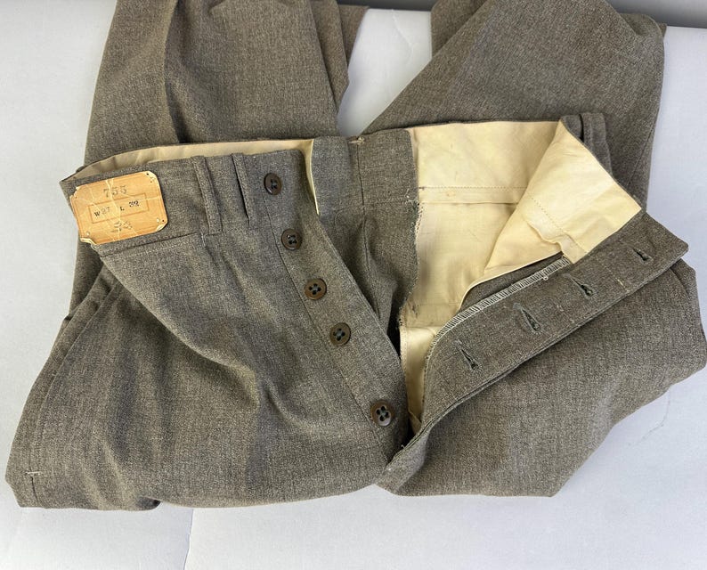 Op de afbeelding: Taupekleurige broek met een knoopsluiting met vijf donkerbruine knopen. De binnenkant van de tailleband is gevoerd met cr&egrave;mekleurige stof. Een rechthoekig beige label aan de linkerkant toont de tekst 755, W 37 L 32.