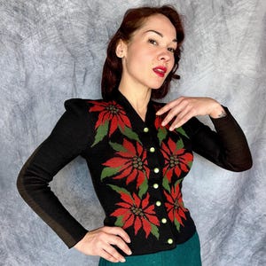May include: Black knit cardigan with red and green floral design. The long-sleeved sweater features a V-neck and is buttoned up the front with gold-colored buttons. The floral pattern is a poinsettia design.