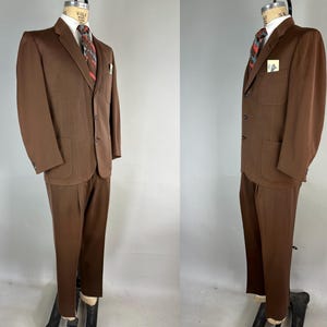 May include: A brown suit displayed on a mannequin. The suit includes a blazer with two front pockets, a matching pair of trousers, a white dress shirt, and a patterned tie. A small card is in the blazer's pocket.