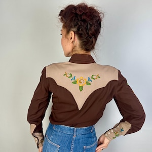 May include: A long-sleeved shirt featuring a dark brown body with a tan yoke on the back. The tan yoke is decorated with an embroidered yellow flower and green leaves. The cuffs are tan with matching floral embroidery and small white buttons.