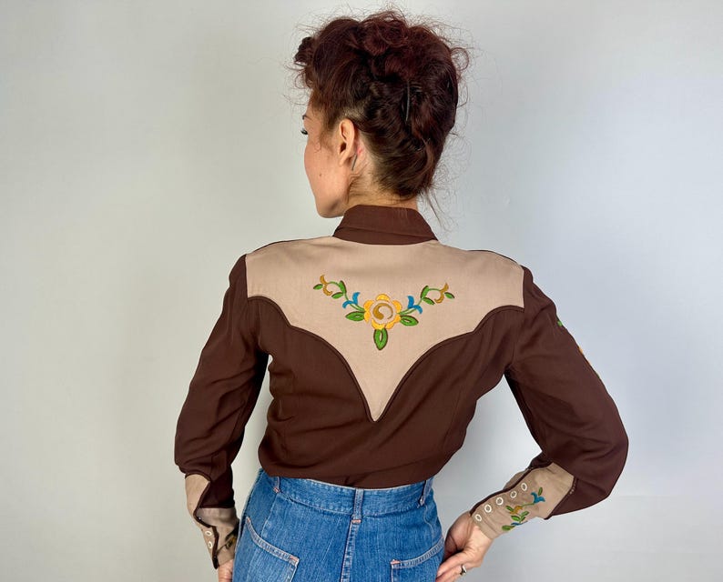 May include: A long-sleeved shirt featuring a dark brown body with a tan yoke on the back. The tan yoke is decorated with an embroidered yellow flower and green leaves. The cuffs are tan with matching floral embroidery and small white buttons.