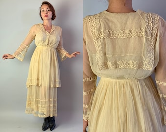 May include: A cream-coloured vintage dress with a tiered skirt and long, sheer sleeves. The dress features delicate lace detailing on the bodice, sleeves, and hem. The dress has a V-neckline and a button-down front. The dress is floor-length.