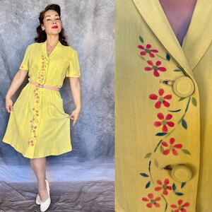 May include: A vintage yellow dress with short sleeves and a button-down front. The dress features a pink belt and floral embroidery. The dress is knee-length and has a classic, retro style.