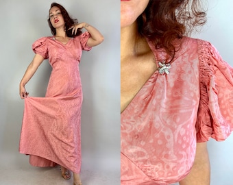May include: A vintage-style, floor-length, rose-colored dress with a subtle floral pattern. The dress features short, puffed sleeves and a V-neckline. Sparkling embellishments adorn the shoulders. The fabric has a slight sheen, adding to its elegant appearance.