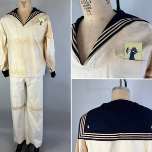 Puede incluir: Un uniforme de marinero blanco vintage con cuello negro y tres rayas doradas. La camisa tiene un pequeño bolsillo con una ilustración de un pájaro. Los pantalones son blancos con pierna ancha.