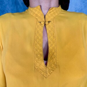 May include: Mustard yellow blouse with a mandarin collar and long sleeves. The collar and front panel feature a quilted diamond pattern and two small, round buttons. The blouse has a keyhole neckline.