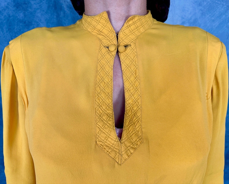 May include: Mustard yellow blouse with a mandarin collar and long sleeves. The collar and front panel feature a quilted diamond pattern and two small, round buttons. The blouse has a keyhole neckline.