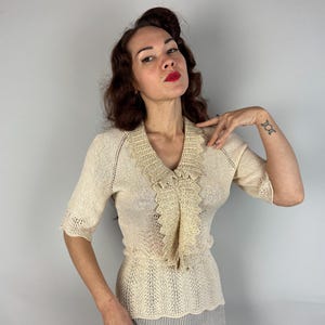 May include: A cream-colored crocheted sweater with a ruffled neckline. The sweater has a short sleeve and a fitted silhouette.