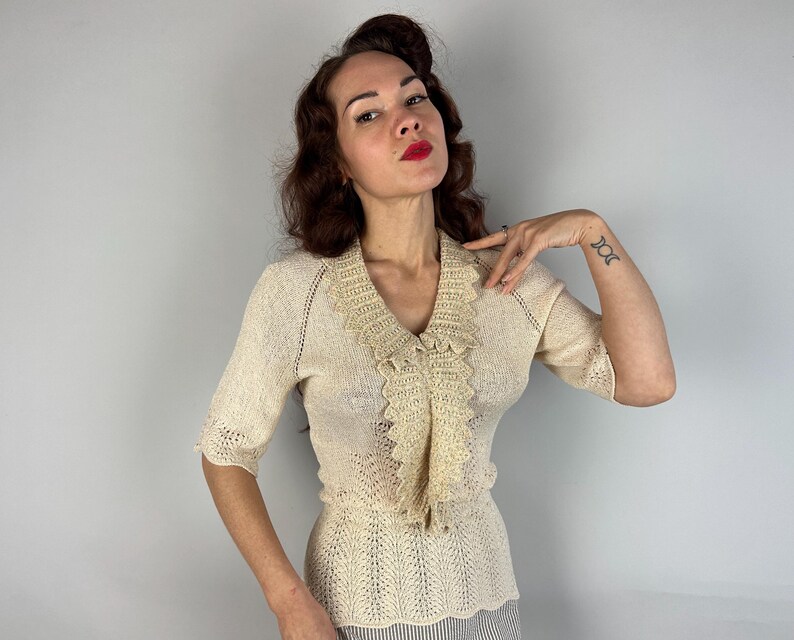 May include: A cream-colored crocheted sweater with a ruffled neckline. The sweater has a short sleeve and a fitted silhouette.