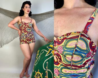 May include: A vintage one-piece swimsuit with a colorful paisley pattern in red, yellow, blue, and pink. The swimsuit has thin straps and a scoop neckline. The model is standing next to a green and yellow nautical-themed umbrella.