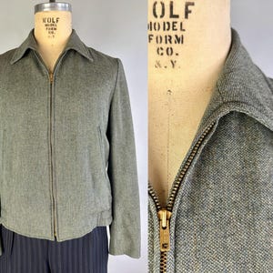 Pode incluir: Um casaco cinzento com fecho de correr feito de tecido texturizado semelhante a lã, com gola pontiaguda e fecho de metal. A peça é exibida num manequim com o texto Wolf Model Co. visível na área do pescoço do busto.