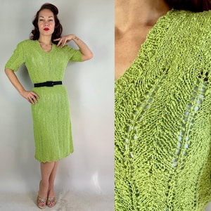 May include: A lime green knitted dress with short sleeves and a V-neck. The dress is cinched at the waist with a black belt. The dress has a textured, open weave design and falls to the mid-calf. The dress is a vintage style.