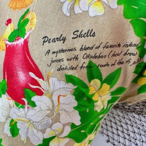 May include: Close-up of a fabric with a tropical theme. A red cocktail with a yellow umbrella and green garnish is surrounded by white and yellow flowers and green leaves. The text "Pearly Shells" is visible.