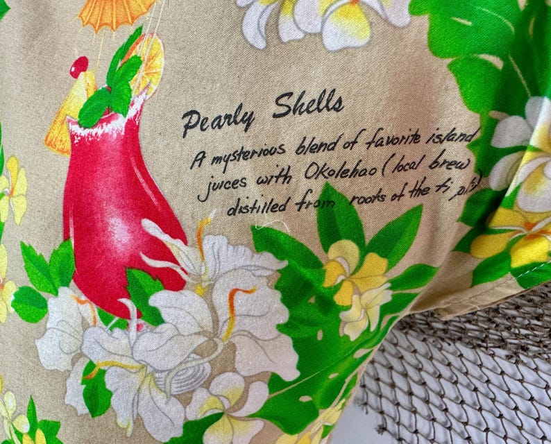 May include: Close-up of a fabric with a tropical theme. A red cocktail with a yellow umbrella and green garnish is surrounded by white and yellow flowers and green leaves. The text "Pearly Shells" is visible.