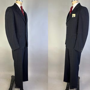 May include: A dark charcoal gray suit consisting of a single-breasted jacket with notched lapels and matching trousers. The jacket features a white collared shirt underneath, a solid red necktie, and a small yellow square pocket square detail.