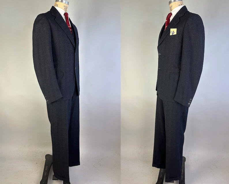 May include: A dark charcoal gray suit consisting of a single-breasted jacket with notched lapels and matching trousers. The jacket features a white collared shirt underneath, a solid red necktie, and a small yellow square pocket square detail.
