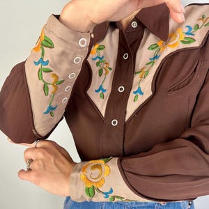 May include: A long-sleeved button-down shirt in dark brown with beige panels on the chest and cuffs. The beige sections feature floral embroidery in yellow, green, and blue. The shirt has a pointed collar and white buttons along the front placket.