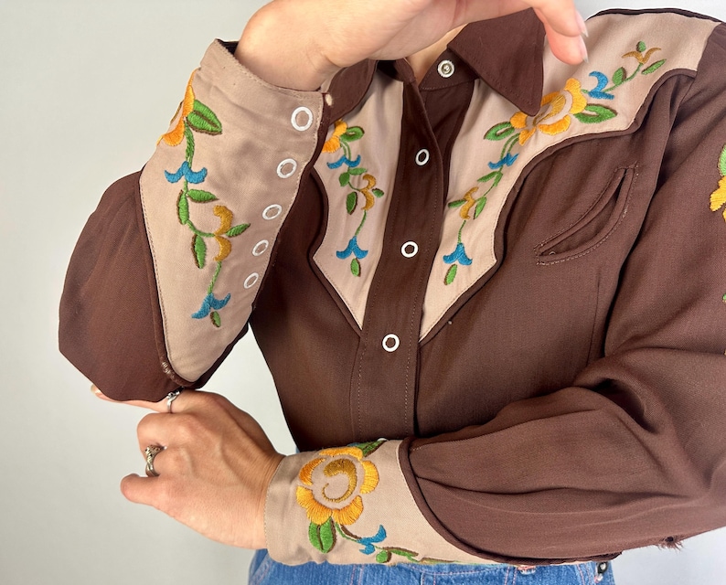 May include: A long-sleeved button-down shirt in dark brown with beige panels on the chest and cuffs. The beige sections feature floral embroidery in yellow, green, and blue. The shirt has a pointed collar and white buttons along the front placket.